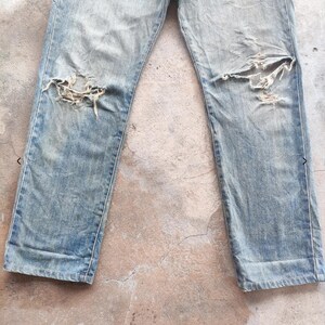 May include: Faded light blue denim jeans with distressed detailing, featuring large rips at the knees. The jeans have a classic five-pocket design and show signs of wear and tear.