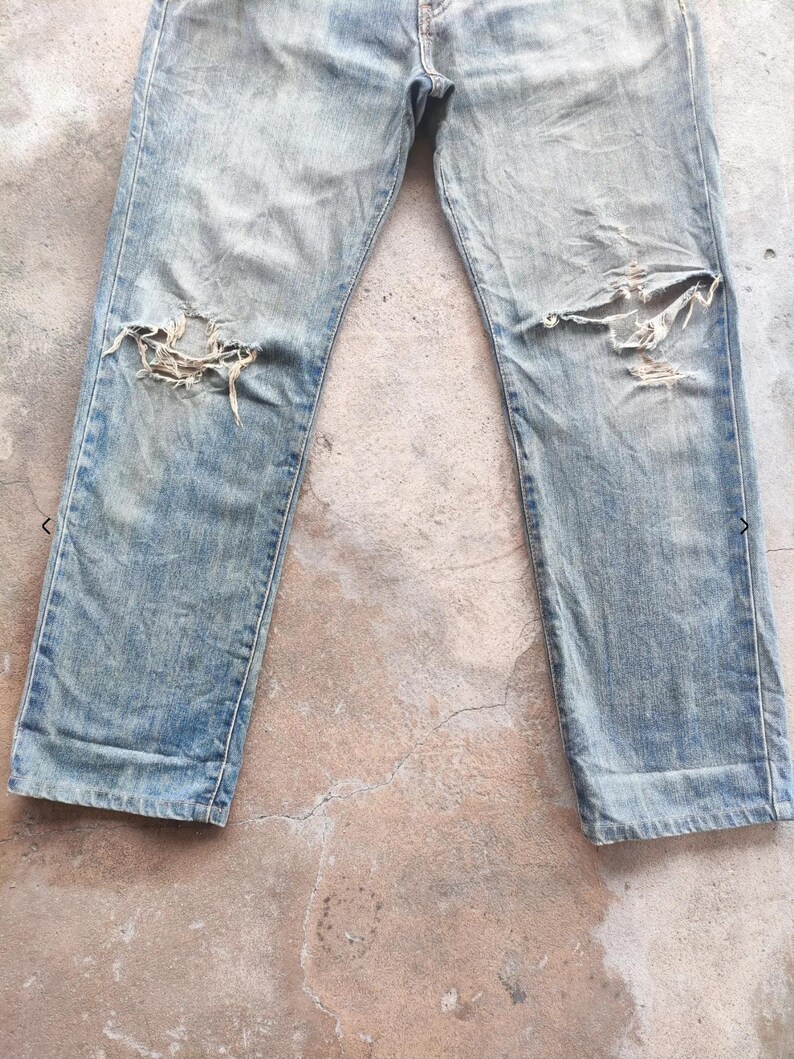 May include: Faded light blue denim jeans with distressed detailing, featuring large rips at the knees. The jeans have a classic five-pocket design and show signs of wear and tear.