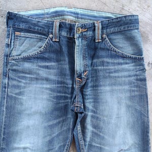 Peut inclure: Gros plan sur un jean en denim bleu d&eacute;lav&eacute;. Le jean pr&eacute;sente une coupe classique &agrave; cinq poches, une fermeture boutonn&eacute;e et des passants de ceinture. La ceinture a un motif ray&eacute;. Le denim a un aspect vieilli avec une d&eacute;coloration et une usure subtiles.
