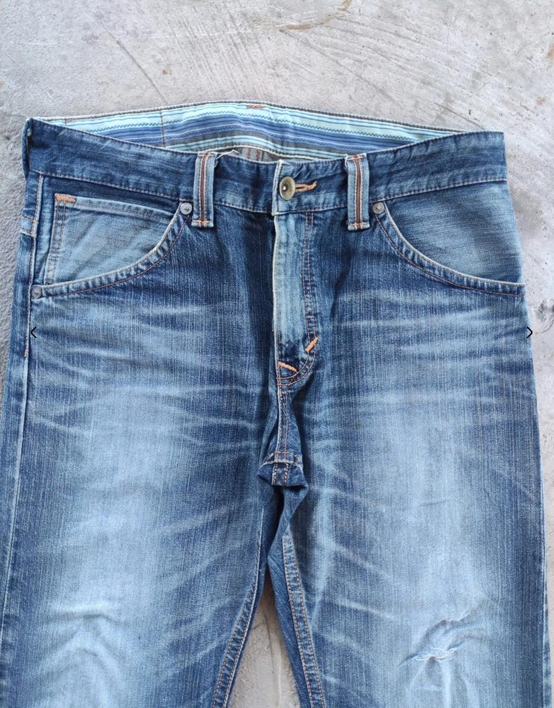 Peut inclure: Gros plan sur un jean en denim bleu d&eacute;lav&eacute;. Le jean pr&eacute;sente une coupe classique &agrave; cinq poches, une fermeture boutonn&eacute;e et des passants de ceinture. La ceinture a un motif ray&eacute;. Le denim a un aspect vieilli avec une d&eacute;coloration et une usure subtiles.