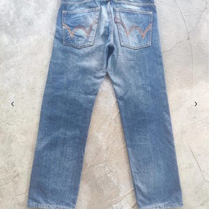 Puede incluir: Vaqueros de mezclilla azul descolorido con un dise&ntilde;o cl&aacute;sico de cinco bolsillos. Cuenta con un parche de cuero marr&oacute;n con la marca "EDWIN" y costuras distintivas en los bolsillos traseros. Los vaqueros tienen un aspecto vintage desgastado.