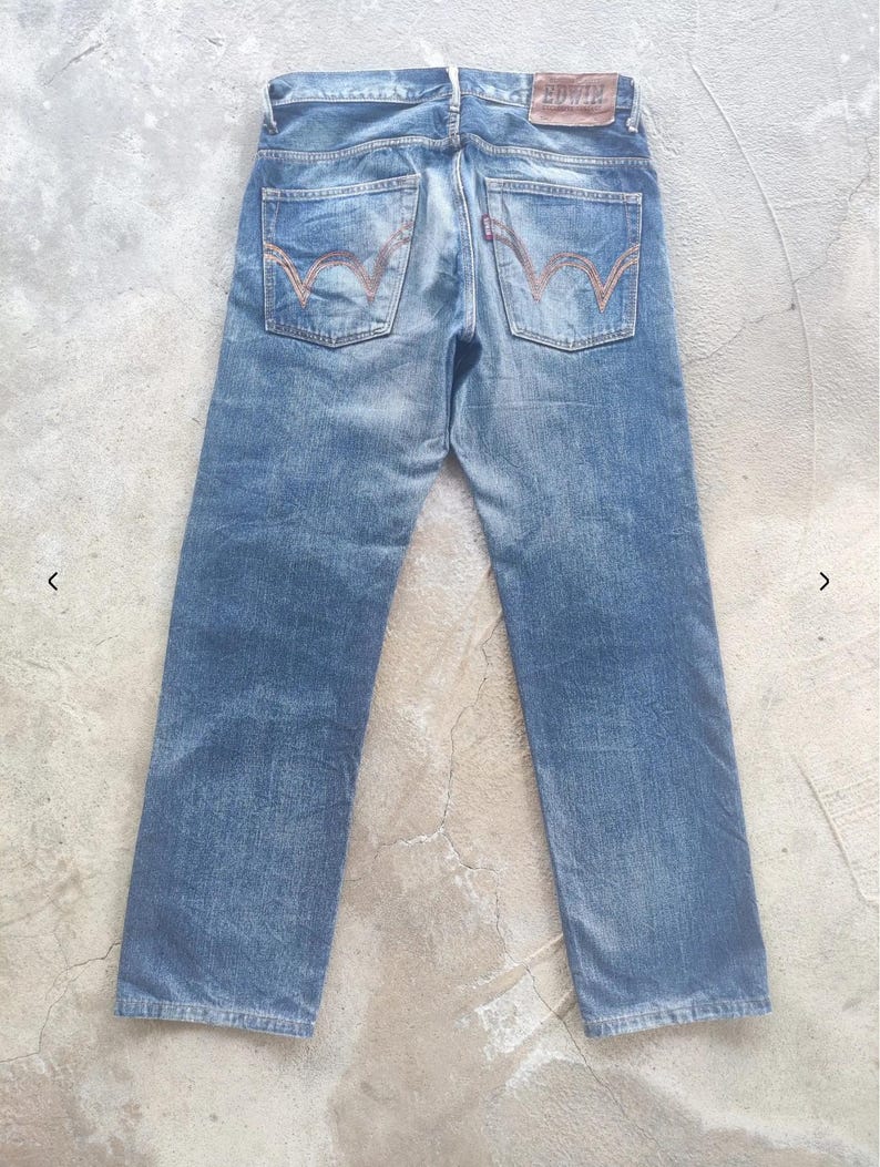 Puede incluir: Vaqueros de mezclilla azul descolorido con un dise&ntilde;o cl&aacute;sico de cinco bolsillos. Cuenta con un parche de cuero marr&oacute;n con la marca "EDWIN" y costuras distintivas en los bolsillos traseros. Los vaqueros tienen un aspecto vintage desgastado.