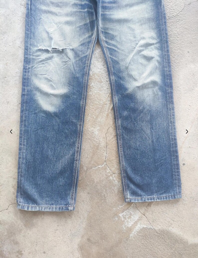 Puede incluir: Vaqueros de mezclilla azul descolorido con un aspecto desgastado. Los vaqueros tienen una rodilla rasgada y marcas de lavado claro. El estilo de pierna recta es visible.