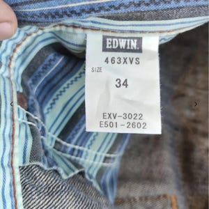 Peut inclure: Gros plan sur une &eacute;tiquette de v&ecirc;tement en jean avec la marque "EDWIN" en blanc sur fond noir. L'&eacute;tiquette affiche la taille "34" et des codes produit. Le v&ecirc;tement a une doublure ray&eacute;e bleue et blanche.