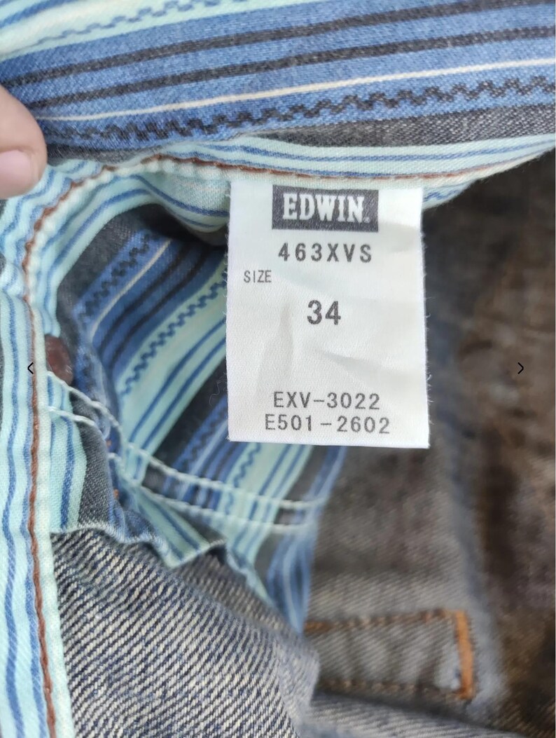 Peut inclure: Gros plan sur une &eacute;tiquette de v&ecirc;tement en jean avec la marque "EDWIN" en blanc sur fond noir. L'&eacute;tiquette affiche la taille "34" et des codes produit. Le v&ecirc;tement a une doublure ray&eacute;e bleue et blanche.