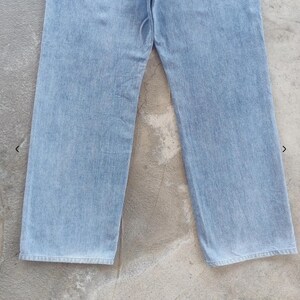 May include: Light blue denim jeans. The jeans have a classic five-pocket design, a straight leg, and a button and zipper closure. The denim has a faded appearance, suggesting a worn-in look. The jeans are laid flat on a concrete surface.