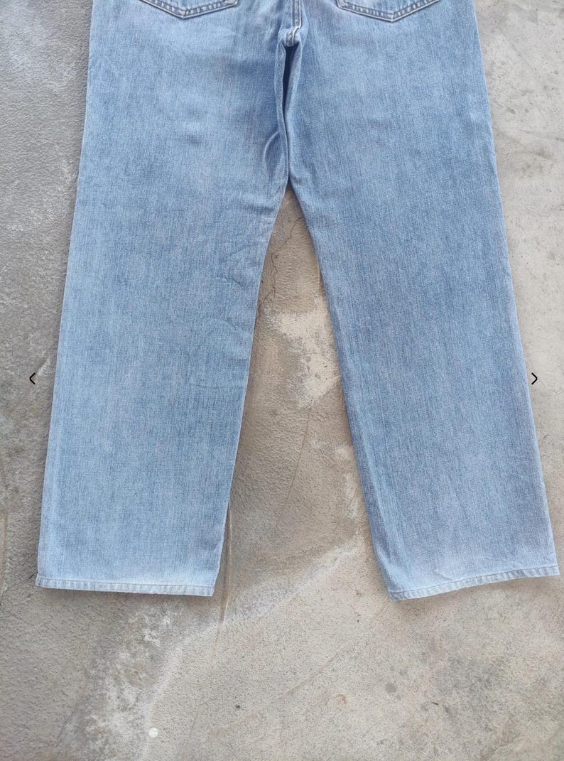 May include: Light blue denim jeans. The jeans have a classic five-pocket design, a straight leg, and a button and zipper closure. The denim has a faded appearance, suggesting a worn-in look. The jeans are laid flat on a concrete surface.