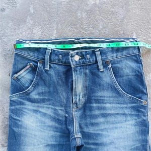 Puede incluir: Primer plano de unos vaqueros azules con una cinta m&eacute;trica en la cintura. Los vaqueros tienen dise&ntilde;os de bolsillo &uacute;nicos y cierre de bot&oacute;n. La cintura mide aproximadamente 35,5 cm.