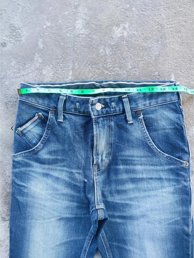 Puede incluir: Primer plano de unos vaqueros azules con una cinta m&eacute;trica en la cintura. Los vaqueros tienen dise&ntilde;os de bolsillo &uacute;nicos y cierre de bot&oacute;n. La cintura mide aproximadamente 35,5 cm.