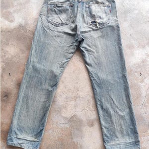 May include: Faded light blue denim jeans with a distressed finish, showing signs of wear and a tear on the back pocket. The jeans have a classic five-pocket design and a brown leather patch.