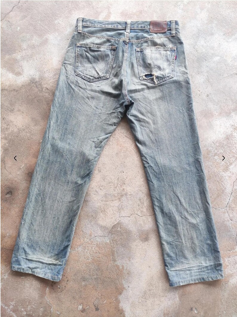 May include: Faded light blue denim jeans with a distressed finish, showing signs of wear and a tear on the back pocket. The jeans have a classic five-pocket design and a brown leather patch.