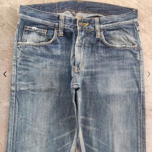 Puede incluir: Vaqueros de mezclilla azul descolorido con un dise&ntilde;o cl&aacute;sico de cinco bolsillos. Los vaqueros tienen cierre de bot&oacute;n y cremallera, trabillas para el cintur&oacute;n y desgaste visible, lo que les da un aspecto vintage. Los vaqueros son de corte recto.