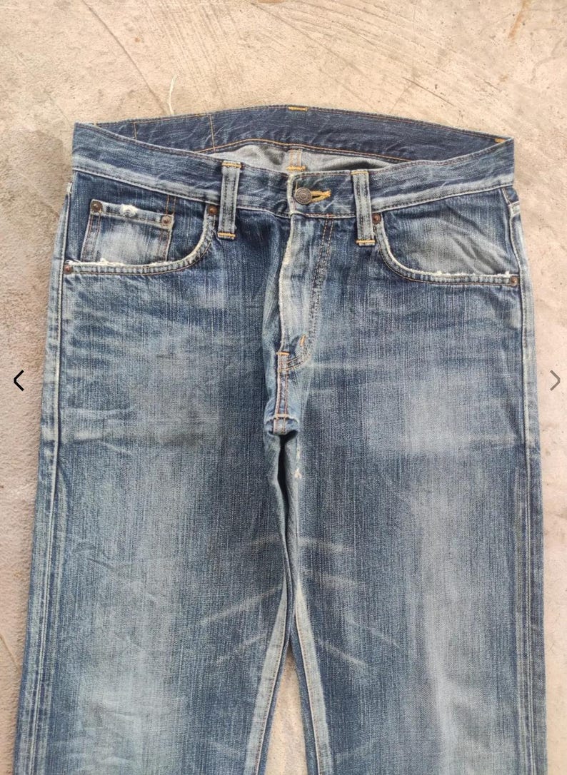 Puede incluir: Vaqueros de mezclilla azul descolorido con un dise&ntilde;o cl&aacute;sico de cinco bolsillos. Los vaqueros tienen cierre de bot&oacute;n y cremallera, trabillas para el cintur&oacute;n y desgaste visible, lo que les da un aspecto vintage. Los vaqueros son de corte recto.
