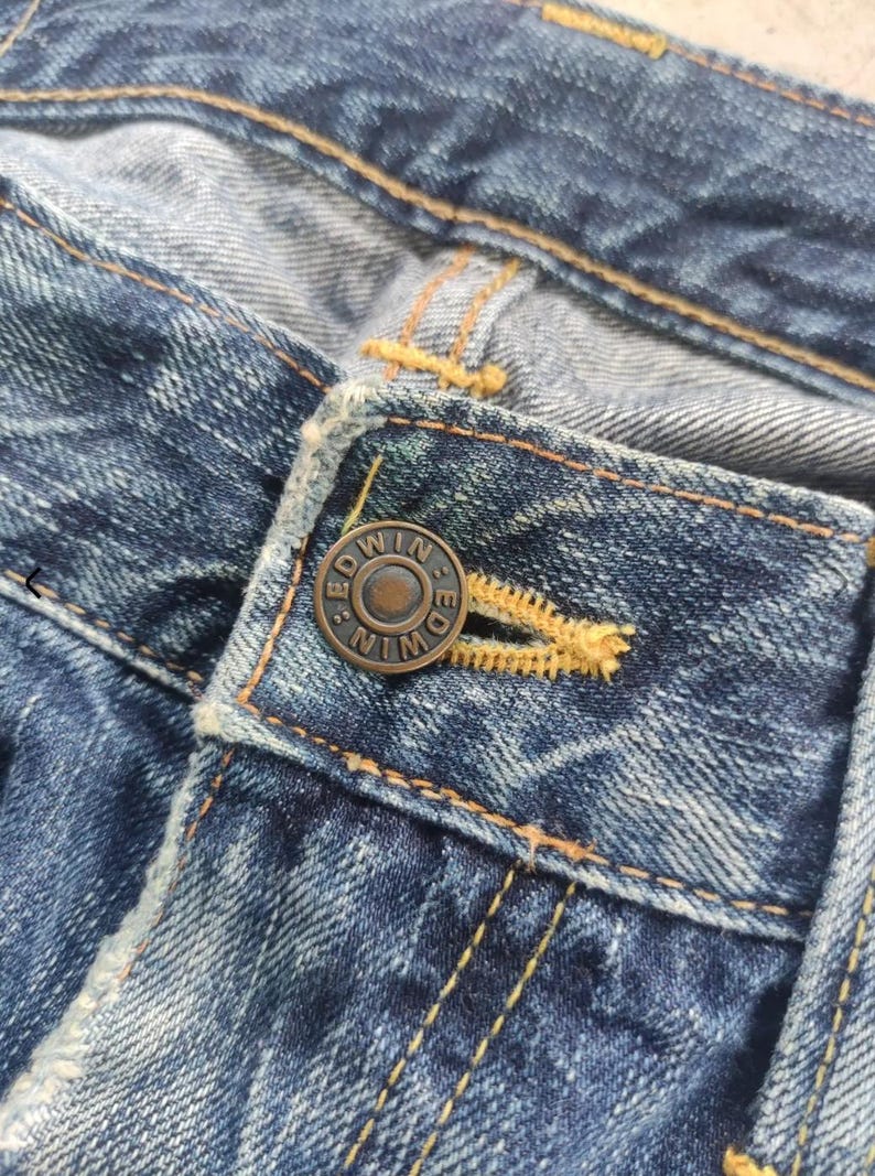 Puede incluir: Primer plano de una secci&oacute;n de vaqueros azules. Los vaqueros presentan un bot&oacute;n met&aacute;lico con la marca "EDWIN" grabada. Las costuras amarillas son visibles a lo largo de las costuras y el ojal, contrastando con la mezclilla.