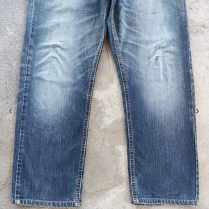 Peut inclure: Jean bleu d&eacute;lav&eacute; avec une conception classique &agrave; cinq poches. Le jean pr&eacute;sente des signes d'usure avec des zones d&eacute;color&eacute;es et une petite d&eacute;chirure sur la jambe droite. La coupe droite et le d&eacute;lavage moyen donnent un aspect vintage.