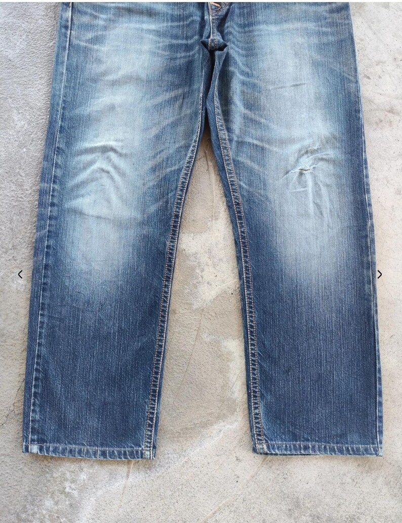 Peut inclure: Jean bleu d&eacute;lav&eacute; avec une conception classique &agrave; cinq poches. Le jean pr&eacute;sente des signes d'usure avec des zones d&eacute;color&eacute;es et une petite d&eacute;chirure sur la jambe droite. La coupe droite et le d&eacute;lavage moyen donnent un aspect vintage.