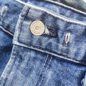 Puede incluir: Primer plano de unos vaqueros azules con un bot&oacute;n y un ojal. El bot&oacute;n plateado lleva la inscripci&oacute;n "LIMITED EDWIN COMPANY". Los vaqueros tienen costuras visibles y un aspecto descolorido.