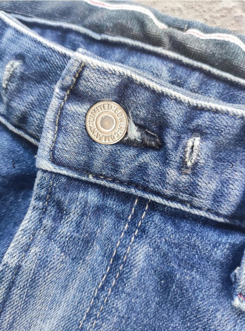 Puede incluir: Primer plano de unos vaqueros azules con un bot&oacute;n y un ojal. El bot&oacute;n plateado lleva la inscripci&oacute;n "LIMITED EDWIN COMPANY". Los vaqueros tienen costuras visibles y un aspecto descolorido.