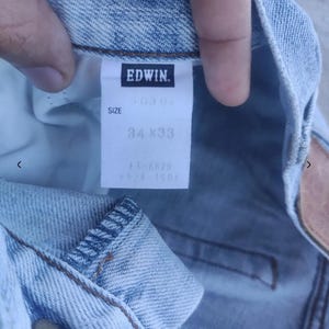May include: Close-up of light blue denim jeans with a tag that reads "EDWIN." The tag also indicates the size as 34x33 inches. The jeans have a classic five-pocket design and a button closure. The denim has a faded, worn look.