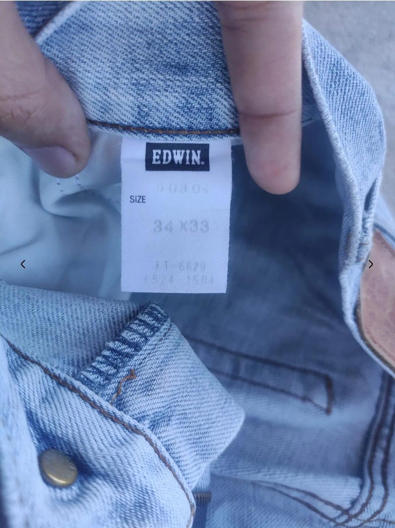 May include: Close-up of light blue denim jeans with a tag that reads "EDWIN." The tag also indicates the size as 34x33 inches. The jeans have a classic five-pocket design and a button closure. The denim has a faded, worn look.
