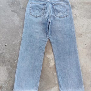 May include: Light blue denim jeans with a classic five-pocket design. The jeans feature a straight leg cut and a brown leather patch on the back waistband. The back pockets have a distinctive curved stitch detail. The jeans are laid flat on a concrete surface.