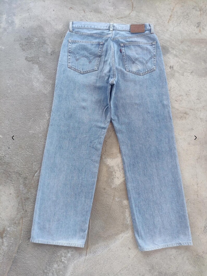 May include: Light blue denim jeans with a classic five-pocket design. The jeans feature a straight leg cut and a brown leather patch on the back waistband. The back pockets have a distinctive curved stitch detail. The jeans are laid flat on a concrete surface.