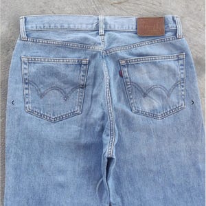 May include: Light blue denim jeans with classic five-pocket styling. Features a brown leather patch with embossed text and the iconic Levi's back pocket stitching. The jeans have a faded wash and a red tab on the back pocket.