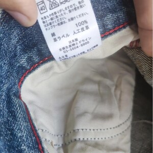 Puede incluir: Primer plano de unos vaqueros de mezclilla con forro interior blanco y costuras rojas. Se ve una etiqueta con texto japon&eacute;s, que incluye instrucciones de lavado y composici&oacute;n de la tela. Los vaqueros son de algod&oacute;n y cuero artificial.