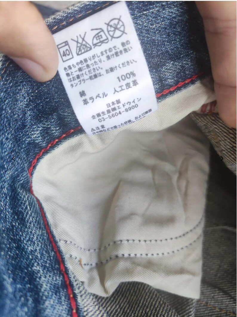 Puede incluir: Primer plano de unos vaqueros de mezclilla con forro interior blanco y costuras rojas. Se ve una etiqueta con texto japon&eacute;s, que incluye instrucciones de lavado y composici&oacute;n de la tela. Los vaqueros son de algod&oacute;n y cuero artificial.