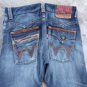 Peut inclure: Jean bleu d&eacute;lav&eacute; avec des d&eacute;tails en cuir marron sur les poches arri&egrave;re. Comprend une poche zipp&eacute;e et une poche boutonn&eacute;e. Le jean porte la marque "EDWIN" sur une &eacute;tiquette en cuir.