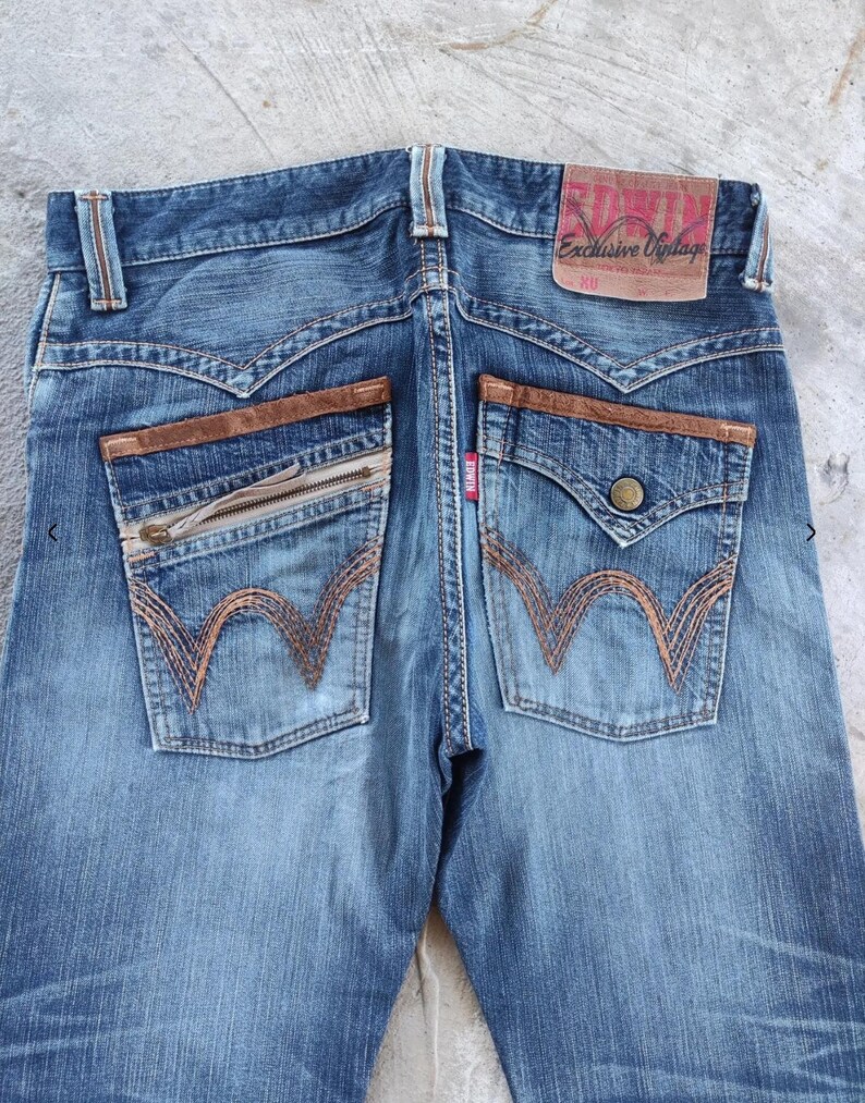 Peut inclure: Jean bleu d&eacute;lav&eacute; avec des d&eacute;tails en cuir marron sur les poches arri&egrave;re. Comprend une poche zipp&eacute;e et une poche boutonn&eacute;e. Le jean porte la marque "EDWIN" sur une &eacute;tiquette en cuir.