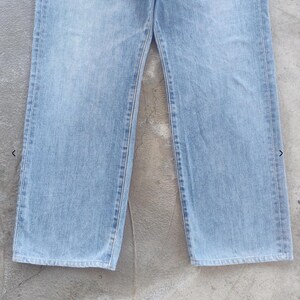May include: Light blue denim jeans. The jeans have a classic five-pocket design, with a straight leg cut. The denim has a faded appearance, with visible stitching along the seams. The jeans are displayed on a gray surface.