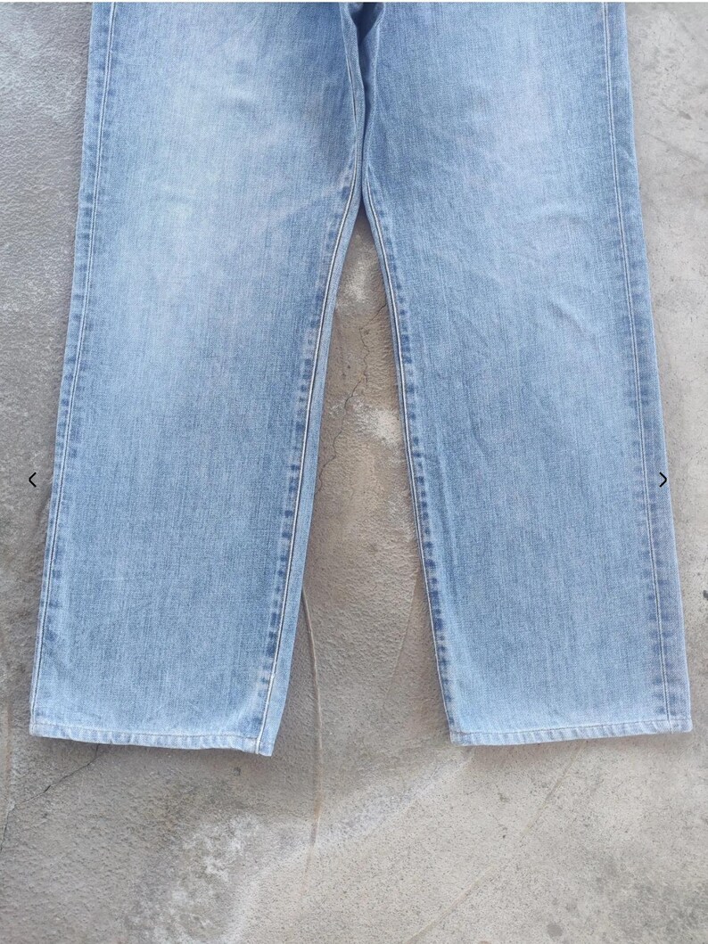 May include: Light blue denim jeans. The jeans have a classic five-pocket design, with a straight leg cut. The denim has a faded appearance, with visible stitching along the seams. The jeans are displayed on a gray surface.