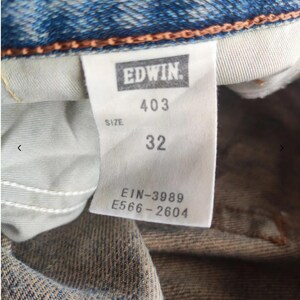May include: Close-up of a blue denim jeans label. The label reads "EDWIN" in a rectangular box, with "403" and "32" below. Additional text includes "SIZE" and codes "EIN-3989" and "E566-2604".