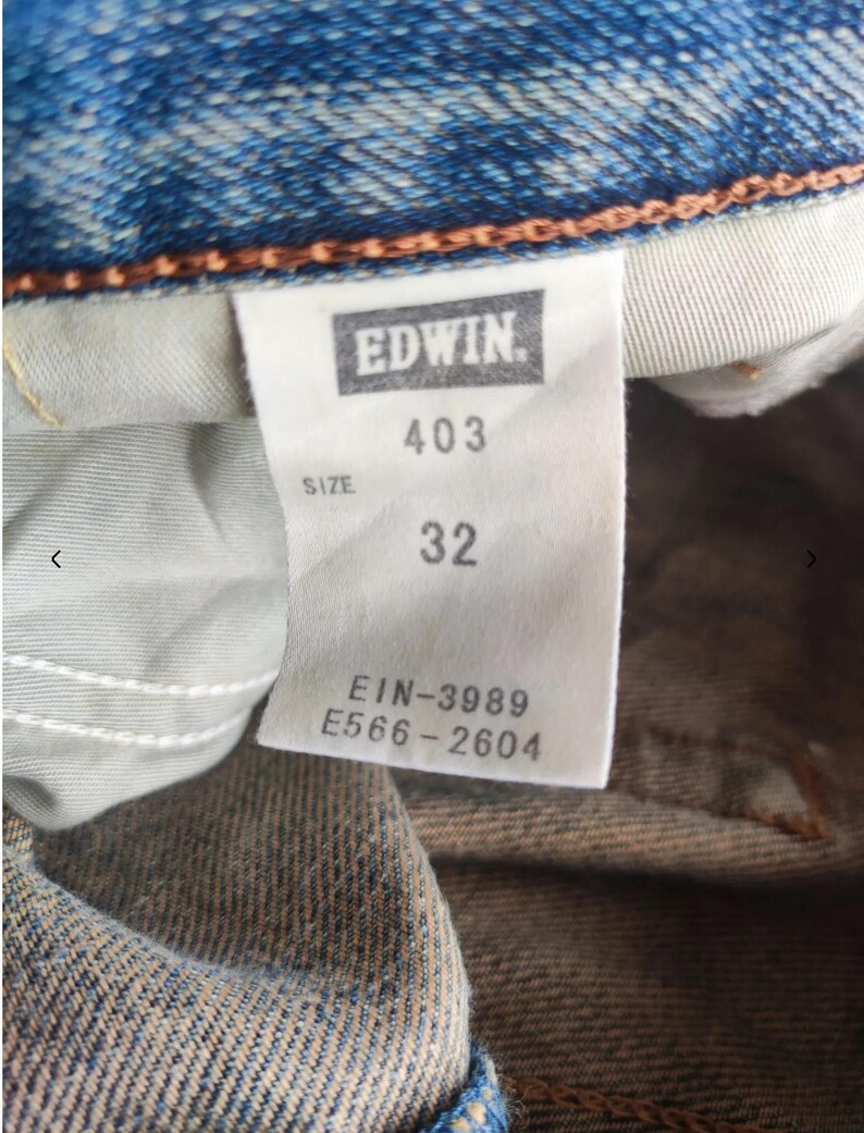 May include: Close-up of a blue denim jeans label. The label reads "EDWIN" in a rectangular box, with "403" and "32" below. Additional text includes "SIZE" and codes "EIN-3989" and "E566-2604".