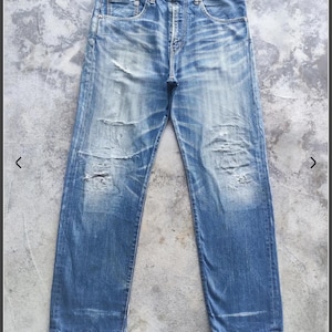 Può includere: Jeans di denim blu sbiadito con dettagli usurati, tra cui strappi e toppe usurate. I jeans hanno un classico design a cinque tasche e una chiusura con bottoni. Il denim ha un lavaggio vintage.