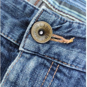 Peut inclure: Gros plan sur un jean en denim bleu avec un bouton en bronze. Le bouton est grav&eacute; de texte et d'&eacute;toiles. Le jean pr&eacute;sente des coutures orange et un d&eacute;tail en tissu ray&eacute; &agrave; la taille.