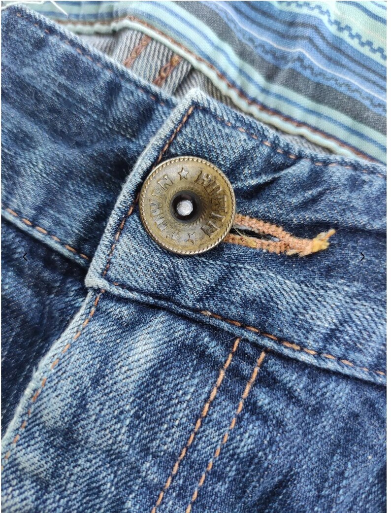 Peut inclure: Gros plan sur un jean en denim bleu avec un bouton en bronze. Le bouton est grav&eacute; de texte et d'&eacute;toiles. Le jean pr&eacute;sente des coutures orange et un d&eacute;tail en tissu ray&eacute; &agrave; la taille.