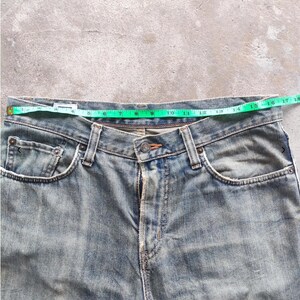 May include: Light blue denim jeans with a green measuring tape across the waist. The jeans have a button closure, pockets, and belt loops. The waist measures approximately 40.6 cm across.