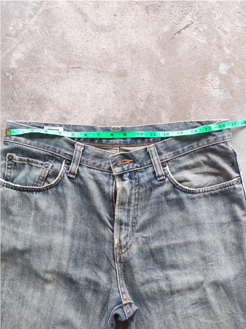 May include: Light blue denim jeans with a green measuring tape across the waist. The jeans have a button closure, pockets, and belt loops. The waist measures approximately 40.6 cm across.