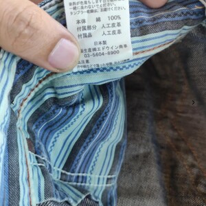 Peut inclure: Gros plan sur un v&ecirc;tement en jean avec une doublure ray&eacute;e bleue et turquoise. Une &eacute;tiquette blanche avec du texte japonais et des symboles d'entretien est visible. L'&eacute;tiquette indique "綿 100%" pour 100% coton.
