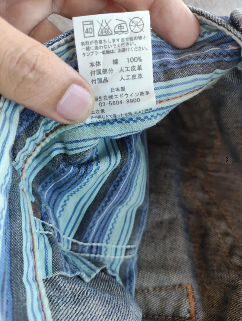 Peut inclure: Gros plan sur un v&ecirc;tement en jean avec une doublure ray&eacute;e bleue et turquoise. Une &eacute;tiquette blanche avec du texte japonais et des symboles d'entretien est visible. L'&eacute;tiquette indique "綿 100%" pour 100% coton.