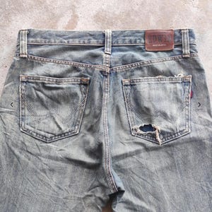 May include: Faded blue denim jeans with distressed details, including a hole in the back pocket. The jeans feature classic five-pocket styling and a brown leather patch with the "EDWIN" brand name. Made in Japan.