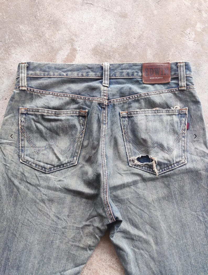 May include: Faded blue denim jeans with distressed details, including a hole in the back pocket. The jeans feature classic five-pocket styling and a brown leather patch with the "EDWIN" brand name. Made in Japan.
