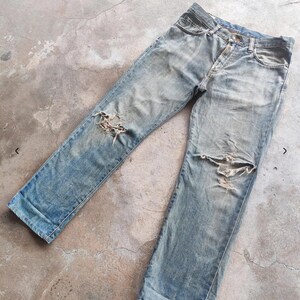 May include: Faded light blue denim jeans with distressed detailing, featuring rips at the knees. The jeans have a classic five-pocket design and a button and zip closure. The fabric shows signs of wear and fading.