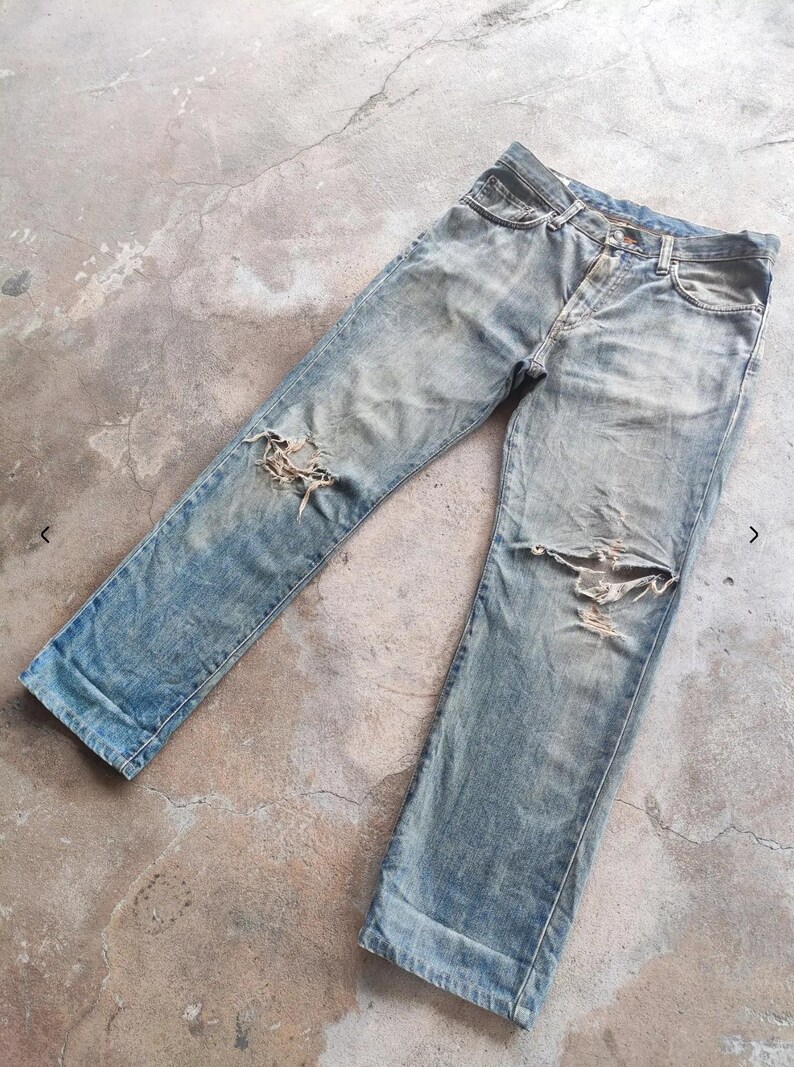 May include: Faded light blue denim jeans with distressed detailing, featuring rips at the knees. The jeans have a classic five-pocket design and a button and zip closure. The fabric shows signs of wear and fading.
