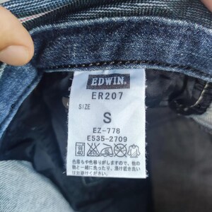 Puede incluir: Primer plano de una etiqueta de prenda vaquera con la marca "EDWIN" y el n&uacute;mero de modelo "ER207". La etiqueta indica la talla "S" e incluye instrucciones de cuidado. La prenda es de mezclilla azul oscuro.
