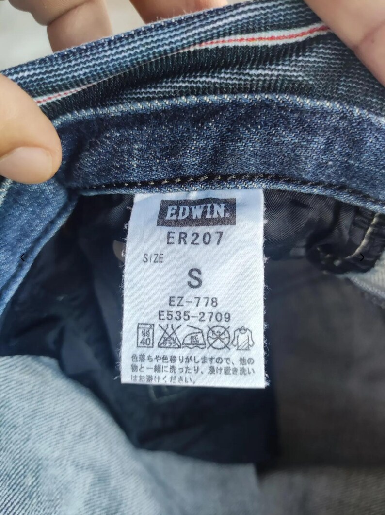 Puede incluir: Primer plano de una etiqueta de prenda vaquera con la marca "EDWIN" y el n&uacute;mero de modelo "ER207". La etiqueta indica la talla "S" e incluye instrucciones de cuidado. La prenda es de mezclilla azul oscuro.