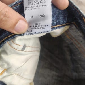 Puede incluir: Primer plano de una etiqueta de jeans de mezclilla azul con texto japon&eacute;s y s&iacute;mbolos de cuidado. La etiqueta indica que los jeans son 100% algod&oacute;n. La etiqueta tambi&eacute;n incluye el nombre y n&uacute;mero de tel&eacute;fono del fabricante.