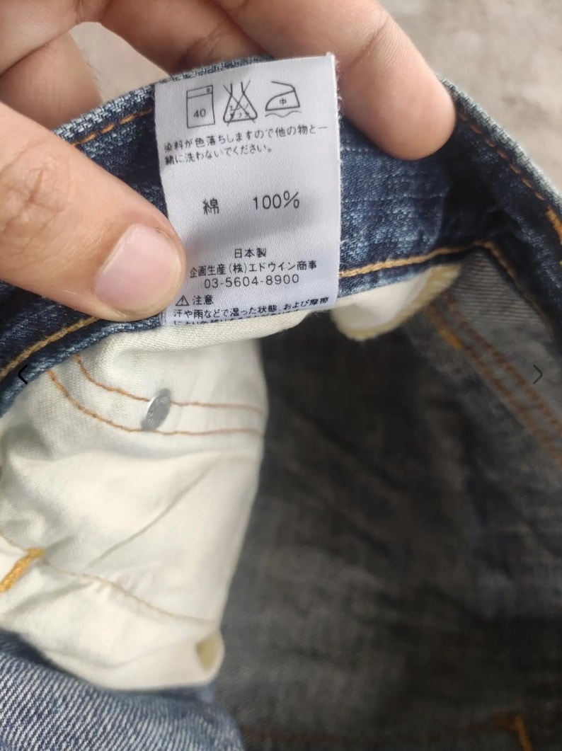 Puede incluir: Primer plano de una etiqueta de jeans de mezclilla azul con texto japon&eacute;s y s&iacute;mbolos de cuidado. La etiqueta indica que los jeans son 100% algod&oacute;n. La etiqueta tambi&eacute;n incluye el nombre y n&uacute;mero de tel&eacute;fono del fabricante.